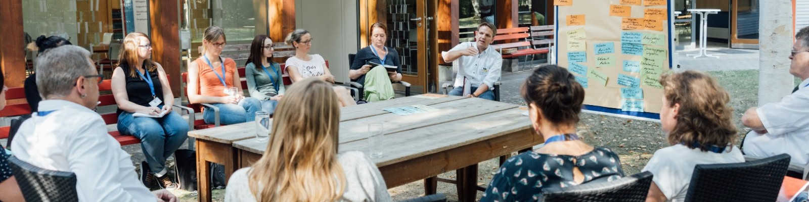 KI generiert: Das Bild zeigt eine Gruppe von Menschen, die im Freien an einem runden Tisch sitzen und sich in einer Diskussion oder einem Meeting befinden. Der Hauptinhalt des Bildes ist eine interaktive Besprechung oder ein Workshop in einer informellen Umgebung.