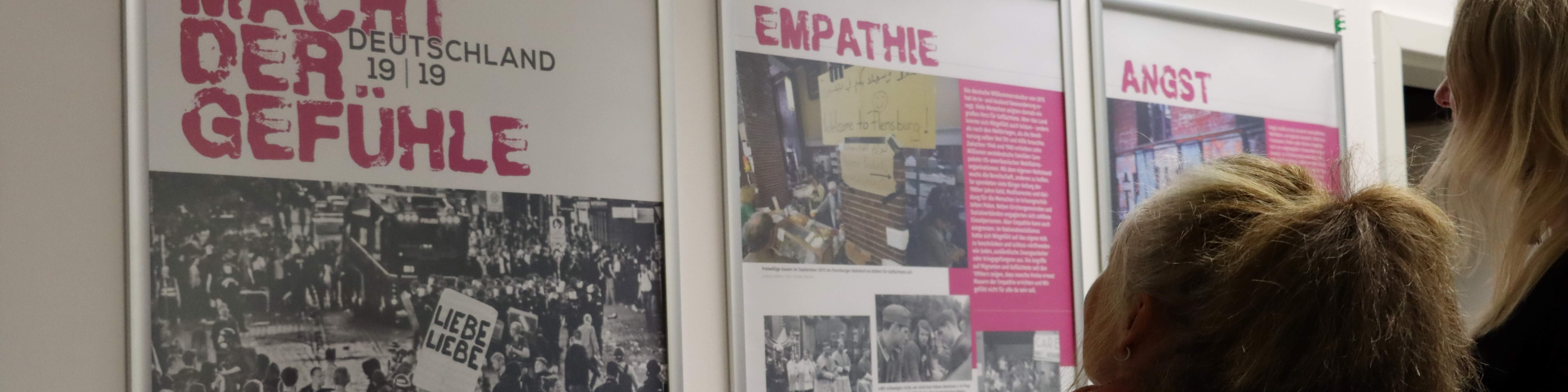 KI generiert: Das Bild zeigt eine Ausstellung mit Postern und Informationen über die Themen "Empathie" und "Angst". Im Vordergrund sind zwei Personen, die sich die Ausstellungsstücke ansehen, während im Hintergrund ein großes Poster mit dem Titel "Macht der Gefühle Deutschland 19 | 19" zu sehen ist.