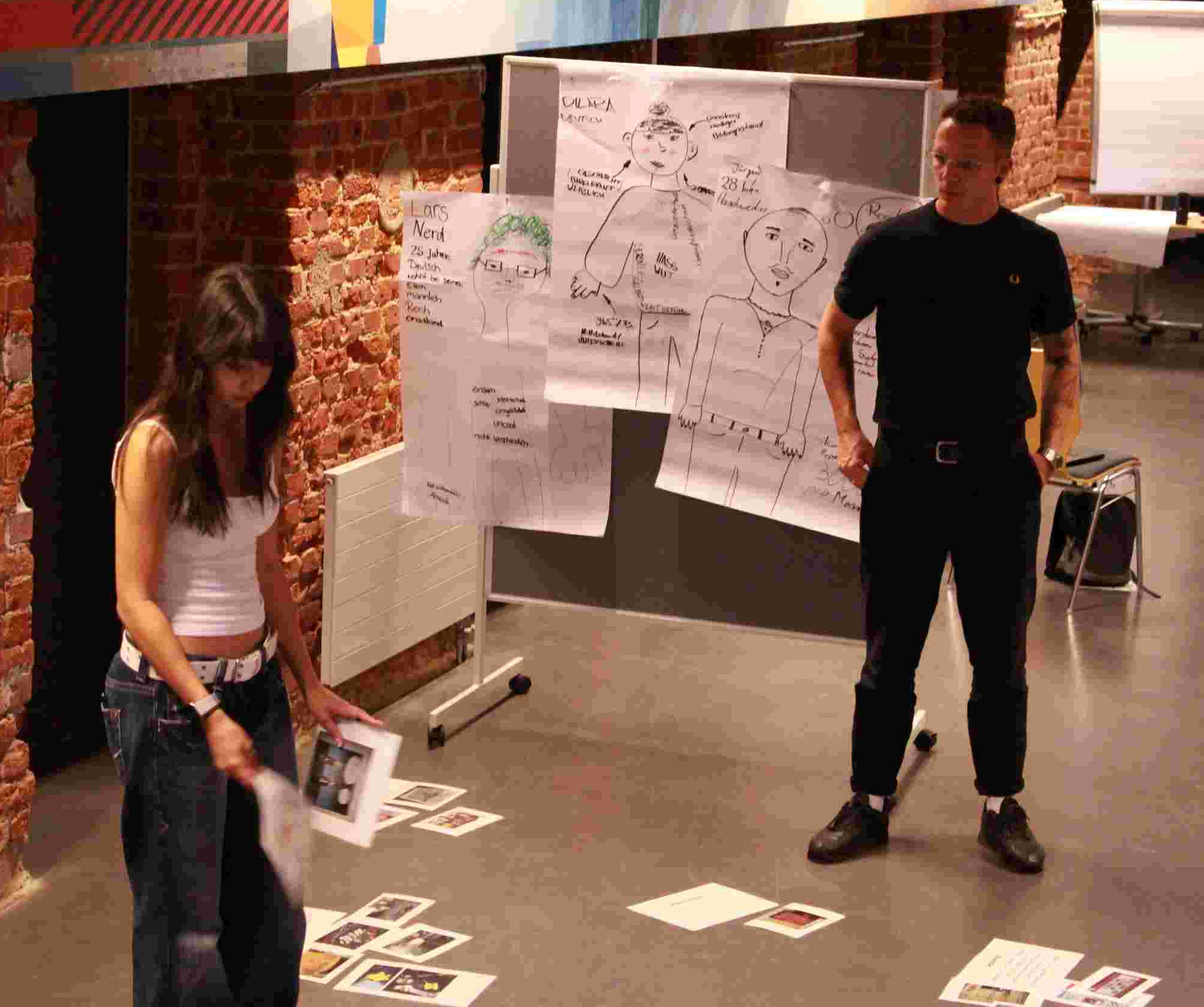 KI generiert: Das Bild zeigt zwei Personen in einem Raum mit Ziegelwänden, die an einer Art Workshop oder Brainstorming-Sitzung teilnehmen. Der Hauptinhalt des Bildes ist die Interaktion der Personen mit den auf dem Boden verstreuten Karten und den an der Wand befestigten Flipcharts mit Notizen und Zeichnungen.