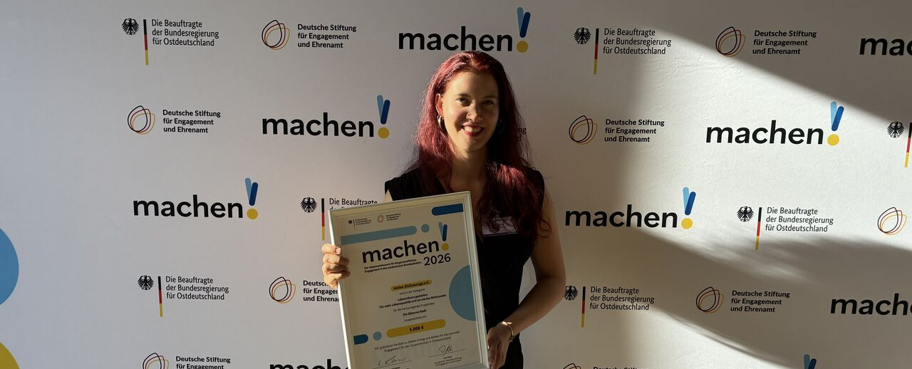 KI generiert: Das Bild zeigt eine Person, die vor einem Step-and-Repeat-Banner mit dem Aufdruck "machen!" und verschiedenen Logos steht und ein Zertifikat oder eine Urkunde mit der Aufschrift "machen!2026" in der Hand hält. Der Hauptinhalt des Bildes ist die Präsentation oder Auszeichnung, die die Person erhalten hat.