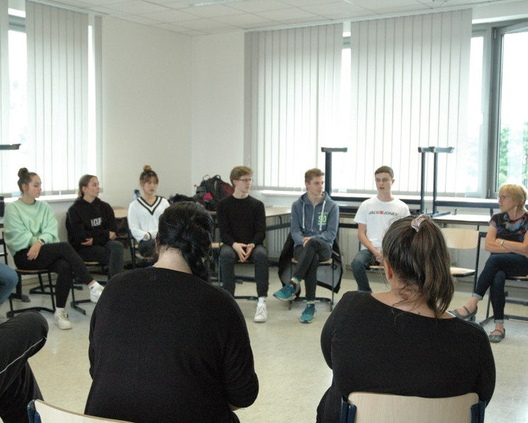 KI generiert: Das Bild zeigt eine Person, die vor einer Gruppe von sitzenden Zuhörern steht und eine Präsentation hält. Im Hintergrund sind Flipcharts mit Notizen und einige bunte Bilder an der Wand zu sehen.