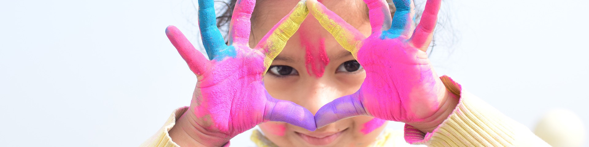 KI generiert: Das Bild zeigt ein Kind mit bunt bemalten Händen, das durch einen Rahmen aus seinen Händen schaut und dabei lächelt. Der Hauptinhalt des Bildes ist die kreative und spielerische Darstellung des Kindes mit den farbenfrohen Handabdrücken.