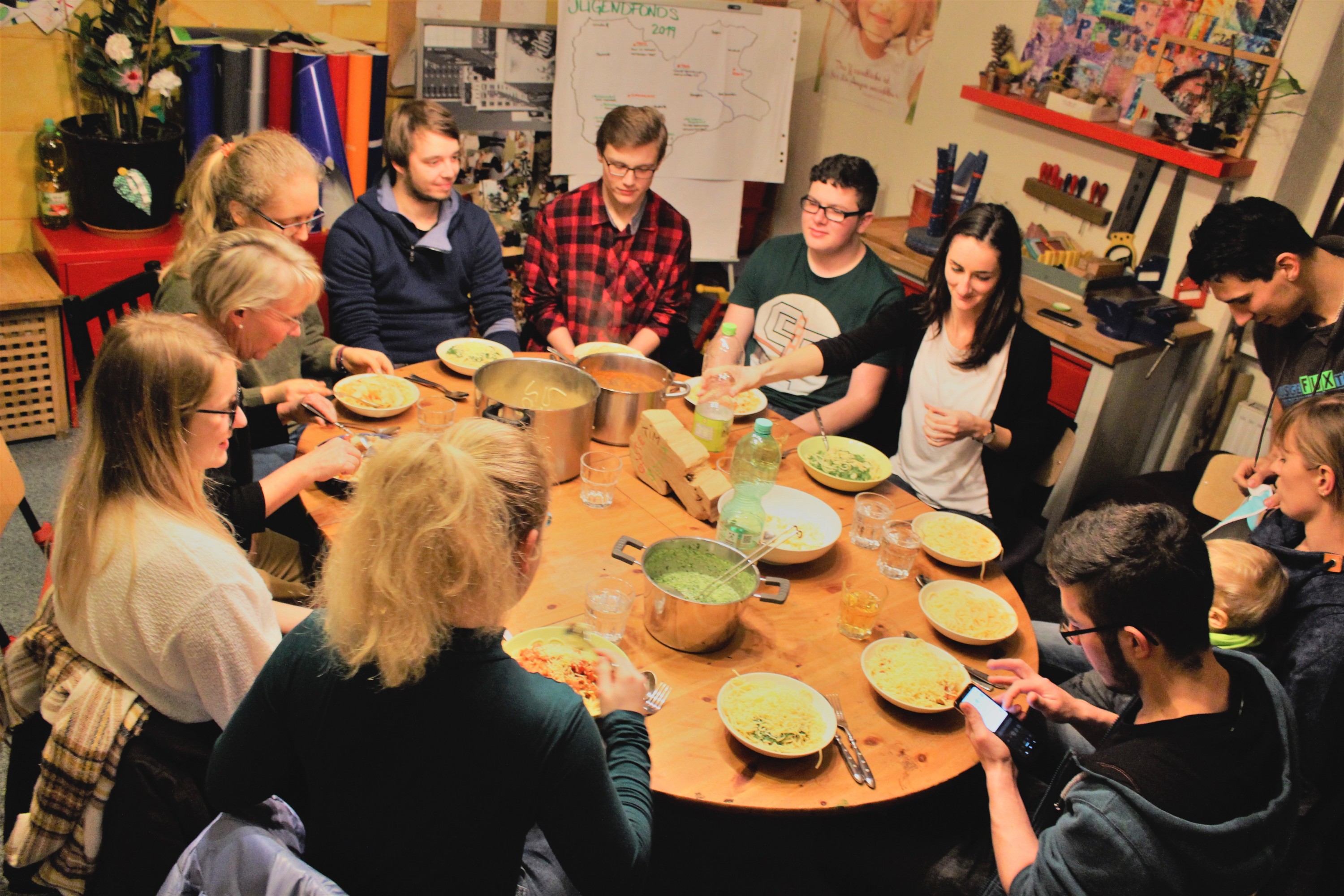 KI generiert: Das Bild zeigt eine Gruppe von Menschen, die gemeinsam an einem großen Holztisch sitzen und verschiedene Gerichte aus großen Töpfen servieren. Der Hauptinhalt des Bildes ist ein geselliges Essen in einer gemütlichen, informellen Atmosphäre.