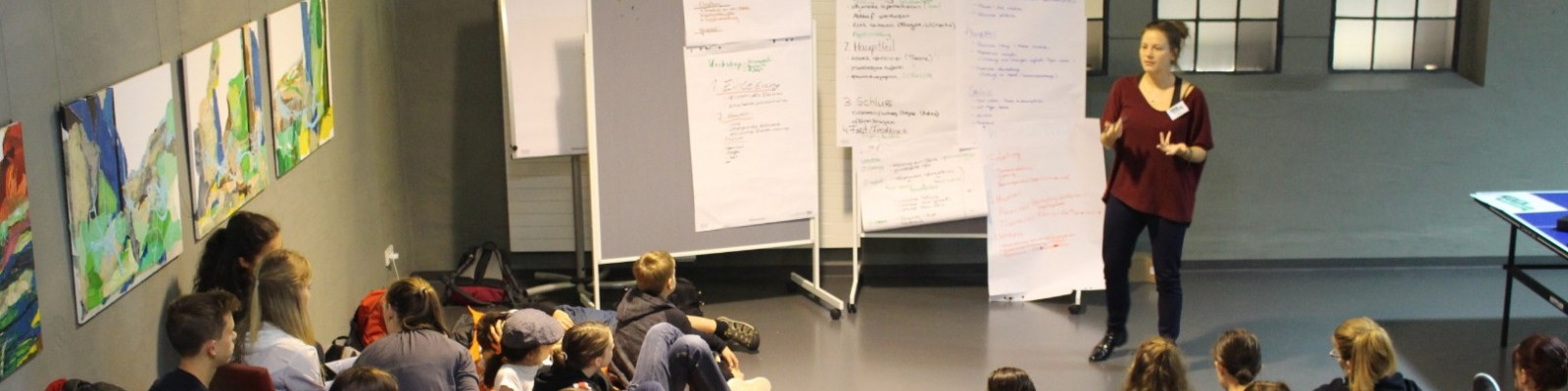 KI generiert: Das Bild zeigt eine Person, die vor einer Gruppe von sitzenden Zuhörern steht und eine Präsentation hält. Im Hintergrund sind Flipcharts mit Notizen und einige bunte Gemälde an der Wand zu sehen.