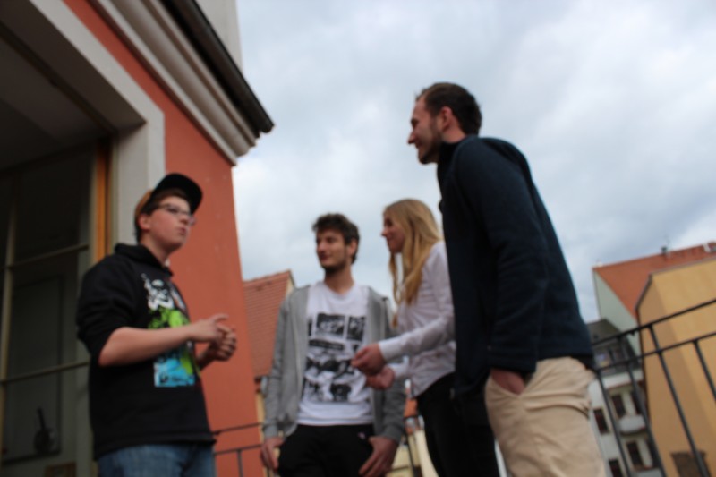 KI generiert: Das Bild zeigt vier Personen, die auf einem Balkon stehen und sich unterhalten. Der Hauptinhalt des Bildes ist die soziale Interaktion zwischen den Personen in einer urbanen Umgebung.