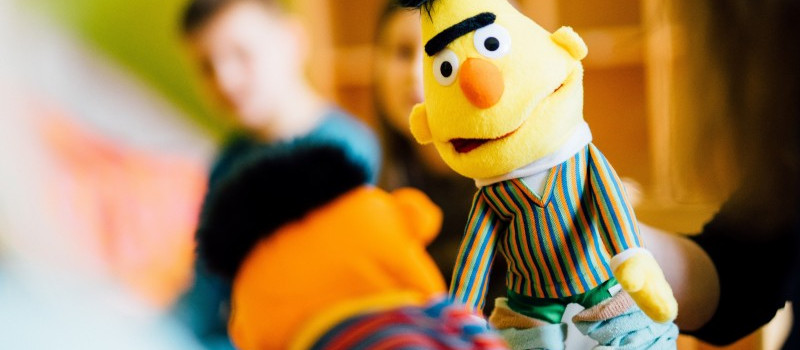 KI generiert: Das Bild zeigt eine Puppe von Bert aus der Sesamstraße im Vordergrund, während im Hintergrund unscharf zwei Personen zu erkennen sind. Der Hauptinhalt des Bildes ist die Puppe von Bert.