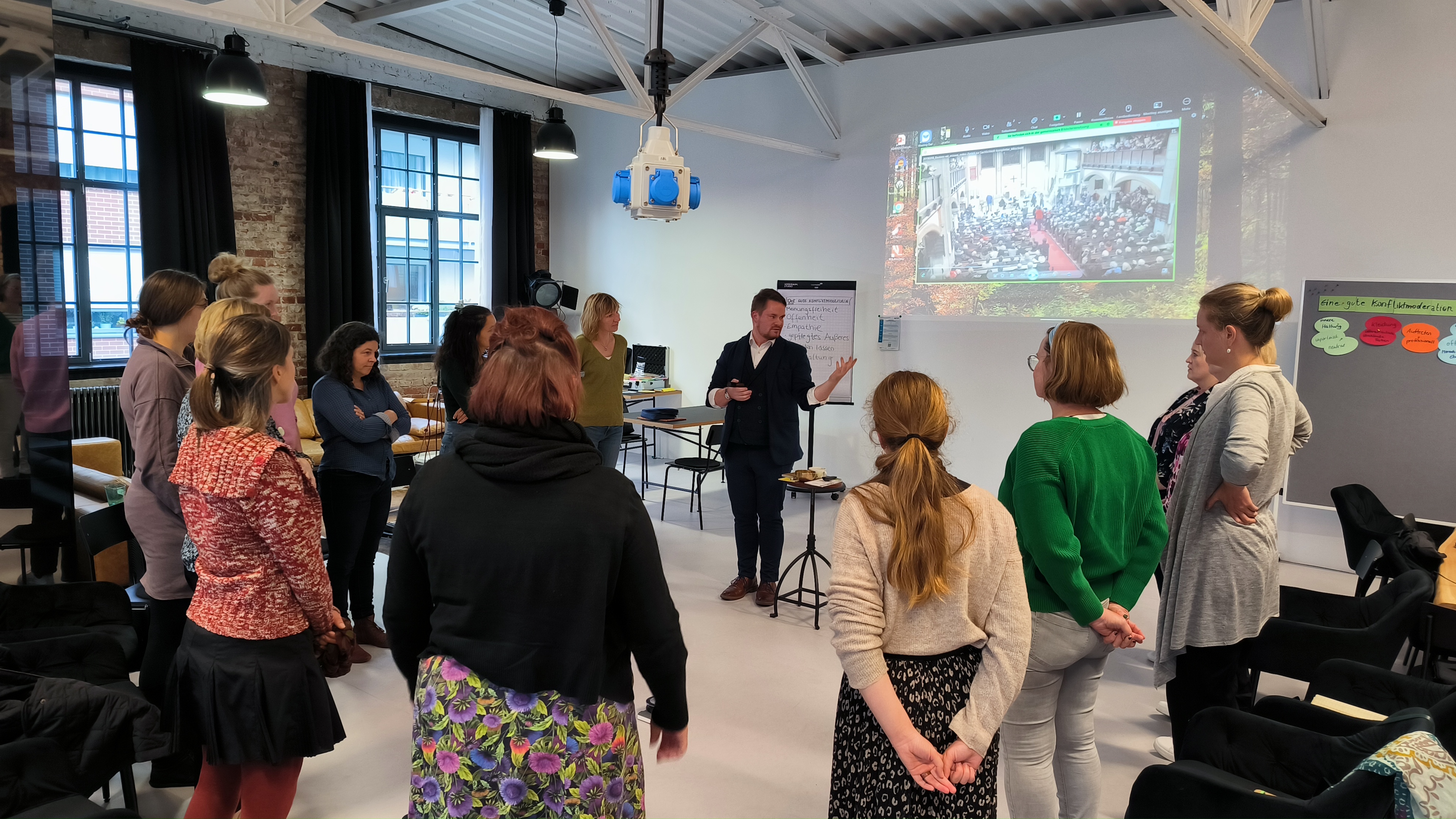 KI generiert: Das Bild zeigt eine Gruppe von Personen in einem hellen Raum, die sich um einen Mann versammelt haben, der vorne steht und eine Präsentation hält. Im Hintergrund ist eine Leinwand mit einer Projektion zu sehen, und die Teilnehmer wirken aufmerksam und engagiert.