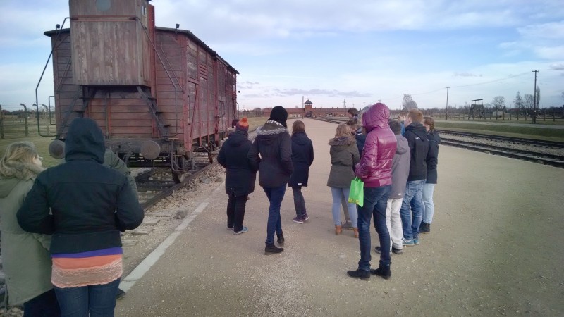 KI generiert: Das Bild zeigt eine Gruppe von Menschen, die sich an einem historischen Ort versammelt haben, wo ein alter Güterwagen auf Gleisen steht. Im Hintergrund ist das Eingangsgebäude eines ehemaligen Konzentrationslagers zu erkennen, was darauf hindeutet, dass es sich um eine Gedenkstätte handelt.