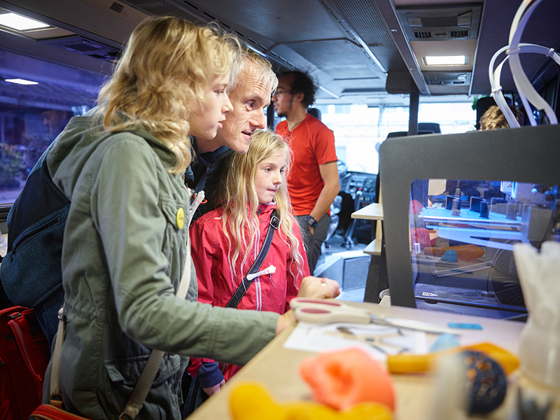 KI generiert: Das Bild zeigt eine Gruppe von Personen, darunter Kinder und Erwachsene, die sich in einem Raum mit technischer Ausstattung befinden und sich für einen 3D-Drucker interessieren. Der Hauptinhalt des Bildes ist die Interaktion und das Lernen über moderne Technologien wie 3D-Druck.