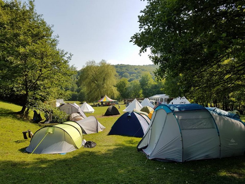KI generiert: Das Bild zeigt einen Campingplatz mit mehreren Zelten, die auf einer grünen Wiese zwischen Bäumen aufgestellt sind. Im Hintergrund sind weitere Zelte und ein großer, weißer Pavillon zu sehen, umgeben von einer bewaldeten Landschaft.