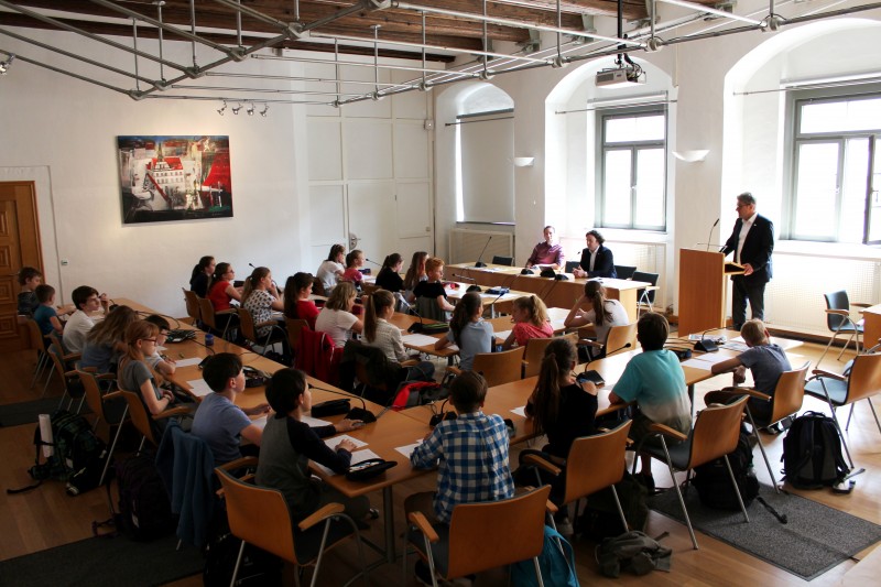KI generiert: Das Bild zeigt eine Gruppe von Schülern in einem Klassenzimmer, die an langen Tischen sitzen und Notizen machen. Vorne steht ein Lehrer oder Redner an einem Pult und spricht zu der Klasse, während zwei weitere Personen an einem Tisch in der Nähe sitzen.
