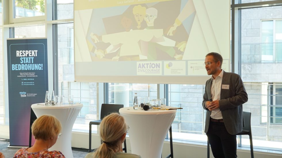 KI generiert: Das Bild zeigt eine Präsentation oder ein Seminar in einem modernen, gut beleuchteten Raum mit großen Fenstern. Ein Mann steht vorne und spricht zu einer kleinen Gruppe von Zuhörern, während auf der Leinwand hinter ihm eine Illustration projiziert wird.