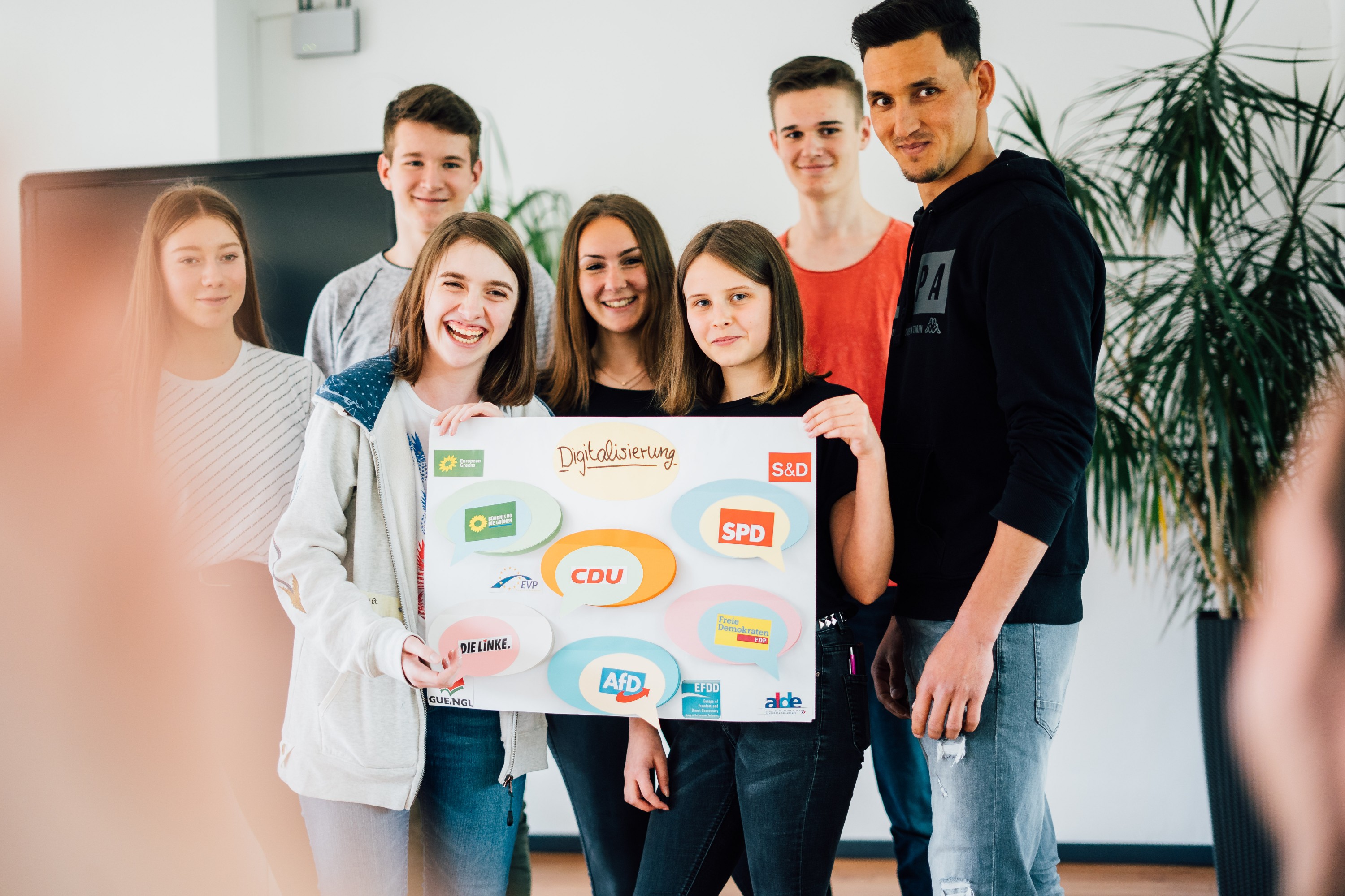 KI generiert: Das Bild zeigt eine Gruppe von sieben Personen, die gemeinsam ein Plakat mit dem Titel "Digitalisierung" und verschiedenen Logos halten. Der Hauptinhalt des Bildes ist die Darstellung einer Teamarbeit oder eines Projekts im Bereich Digitalisierung.