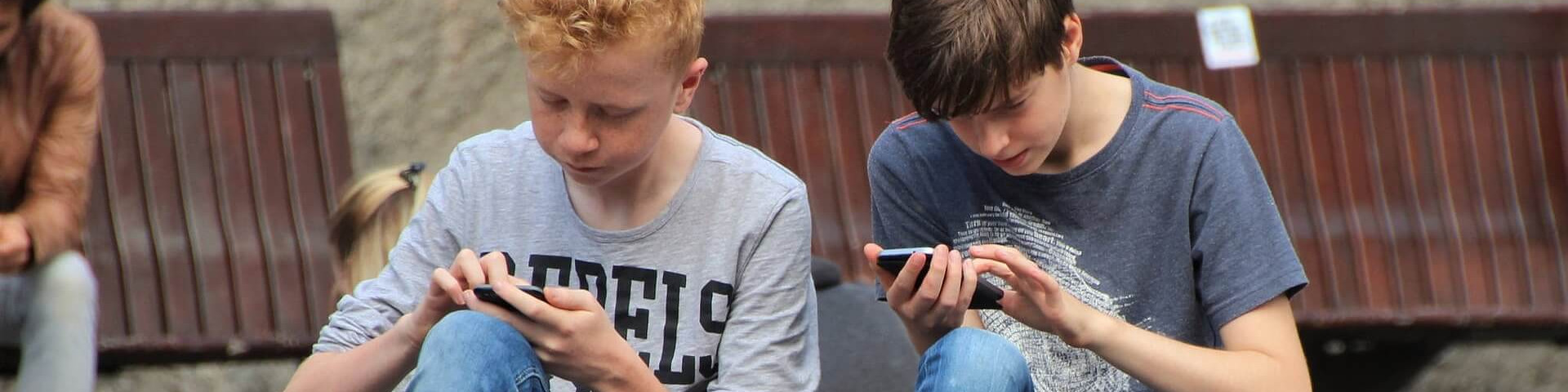 KI generiert: Das Bild zeigt zwei Jugendliche, die auf einer Bank sitzen und intensiv auf ihre Smartphones schauen. Der Hauptinhalt des Bildes ist die Nutzung von Mobiltelefonen durch die Jugendlichen.