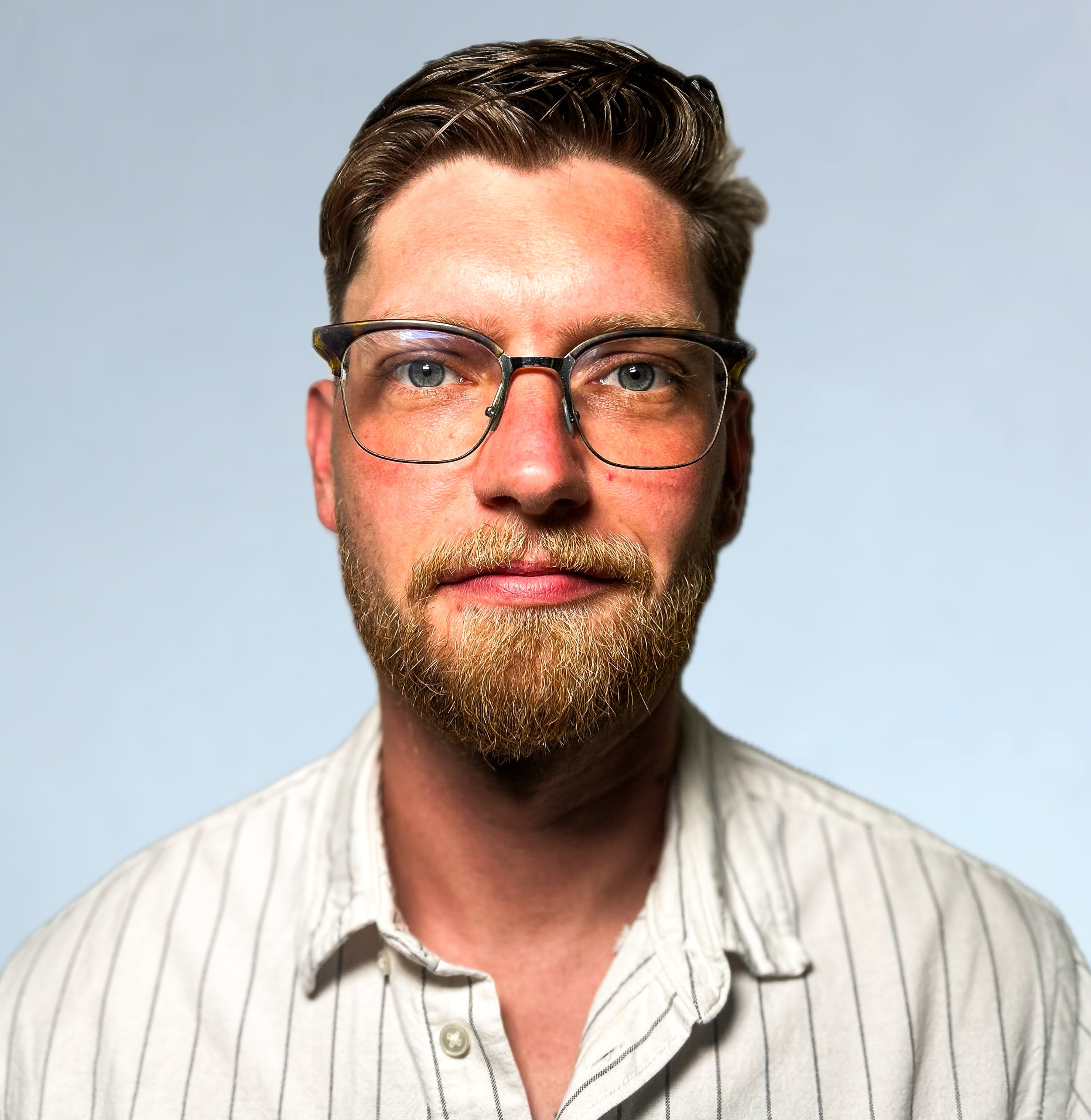 KI generiert: Das Bild zeigt einen Mann mit Brille und Bart, der direkt in die Kamera blickt. Er trägt ein helles, gestreiftes Hemd und hat kurze, braune Haare.