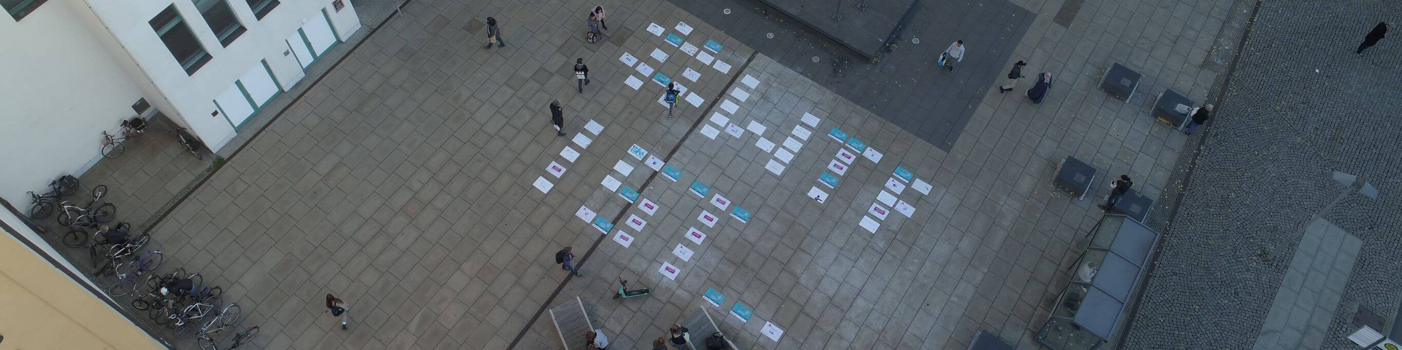 KI generiert: Das Bild zeigt eine Gruppe von Menschen, die auf einem gepflasterten Platz stehen und sich um große, auf dem Boden liegende weiße Blätter mit blauen Aufdrucken versammeln. Der Hauptinhalt des Bildes scheint eine Art Outdoor-Aktivität oder Workshop zu sein, bei dem die Teilnehmer möglicherweise an einer Gruppenarbeit oder einem Spiel teilnehmen.