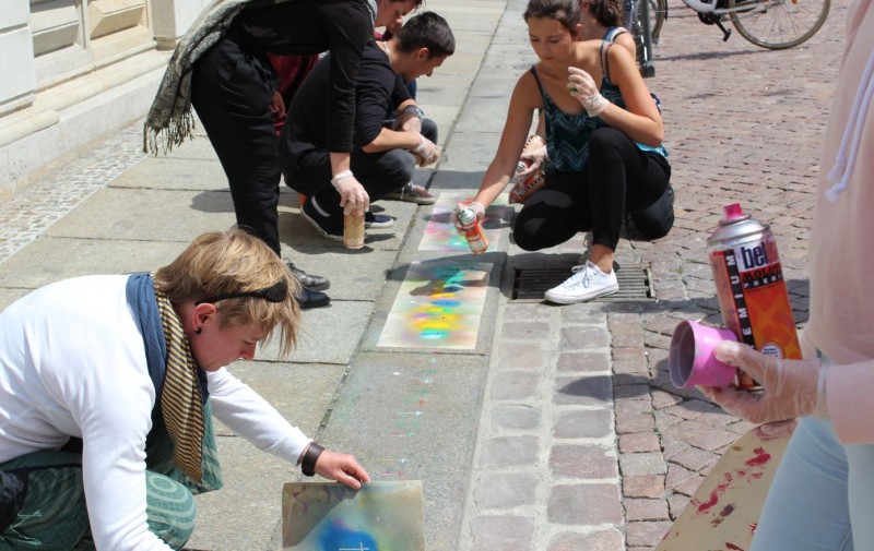 KI generiert: Das Bild zeigt eine Gruppe von Menschen, die auf einem Gehweg mit Sprühdosen und anderen Werkzeugen Graffiti oder Street-Art gestalten. Der Hauptinhalt des Bildes ist die kreative Tätigkeit der Gruppe, die gemeinsam an ihren Kunstwerken arbeitet.