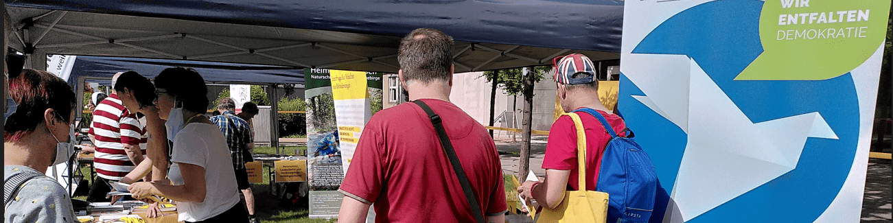 KI generiert: Das Bild zeigt eine Gruppe von Menschen, die sich an einem Stand auf einer Veranstaltung im Freien versammeln. Der Stand hat ein Banner mit der Aufschrift "Wir entfalten Demokratie" und bietet Informationsmaterial an.