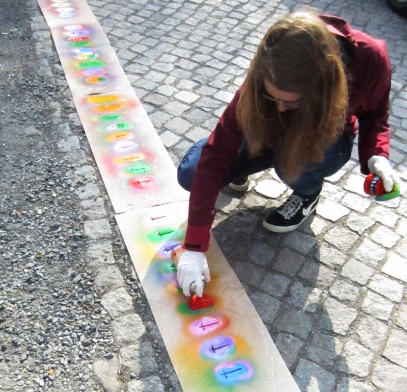 KI generiert: Das Bild zeigt eine Person, die auf einem Gehweg mit Sprühdosen bunte Graffiti auf eine lange Papierbahn sprüht. Der Hauptinhalt des Bildes ist das Graffiti-Sprühen als künstlerische Aktivität.