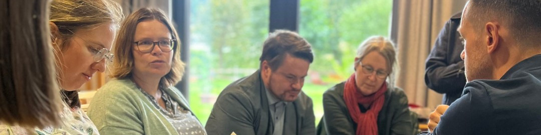 KI generiert: Das Bild zeigt eine Gruppe von fünf Personen, die an einem Tisch sitzen und sich in einer Diskussion oder einem Meeting befinden. Der Hauptinhalt des Bildes ist eine Besprechung oder ein Workshop in einem hellen Raum mit großen Fenstern und Blick auf einen Garten.