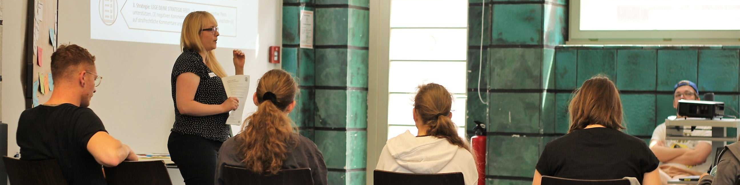 KI generiert: Das Bild zeigt eine Frau, die vor einer kleinen Gruppe von Personen steht und eine Präsentation hält. Der Hauptinhalt des Bildes ist eine Schulungs- oder Seminar-Situation, in der die Referentin vor einem Whiteboard spricht und die Zuhörer aufmerksam zuhören.