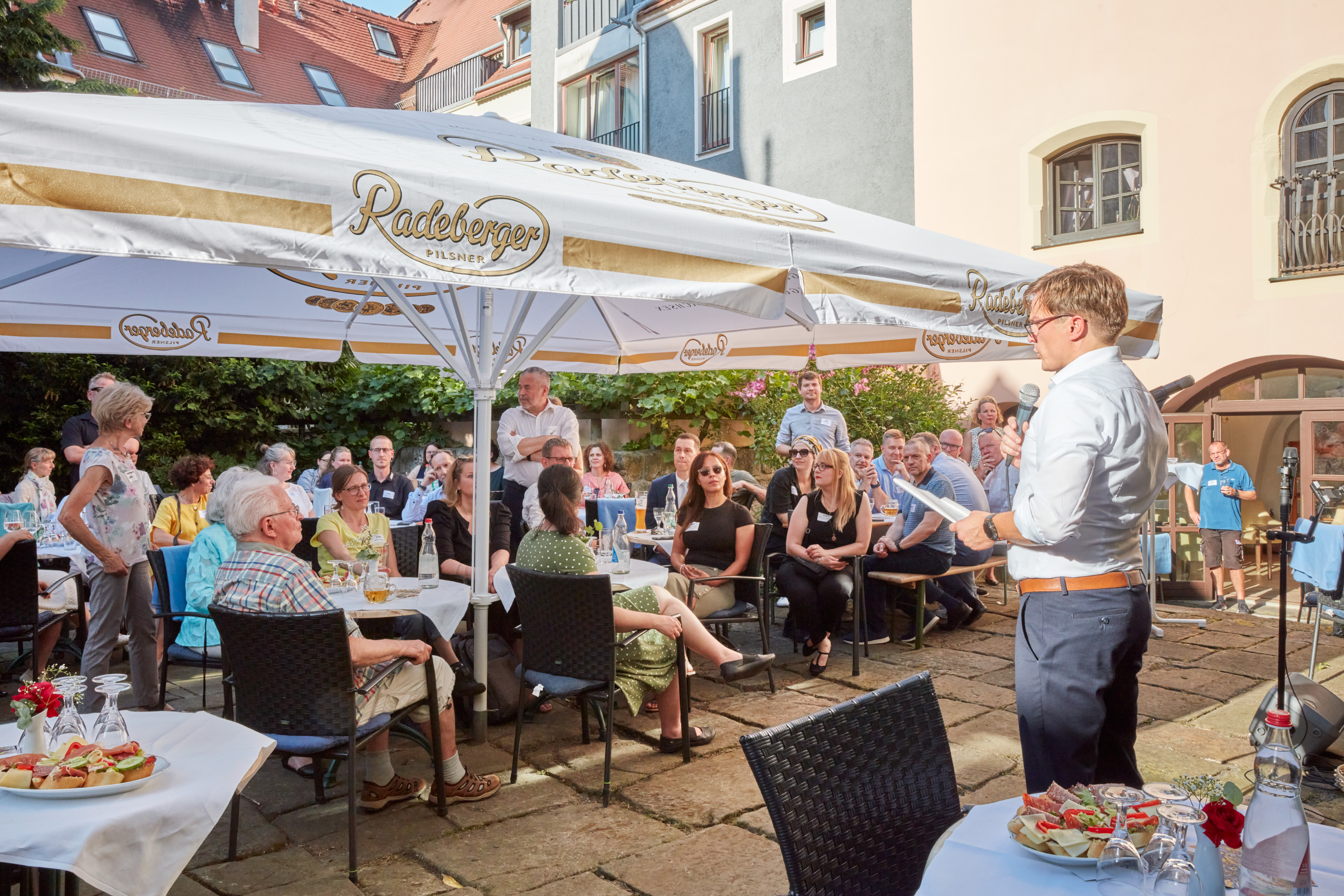 KI generiert: Das Bild zeigt eine Gruppe von Menschen, die im Freien unter einem Radeberger-Bier-Zelt sitzen und einem Mann zuhören, der vor ihnen steht und spricht. Es scheint sich um eine informelle Veranstaltung oder Versammlung in einem Innenhof oder Garten zu handeln.