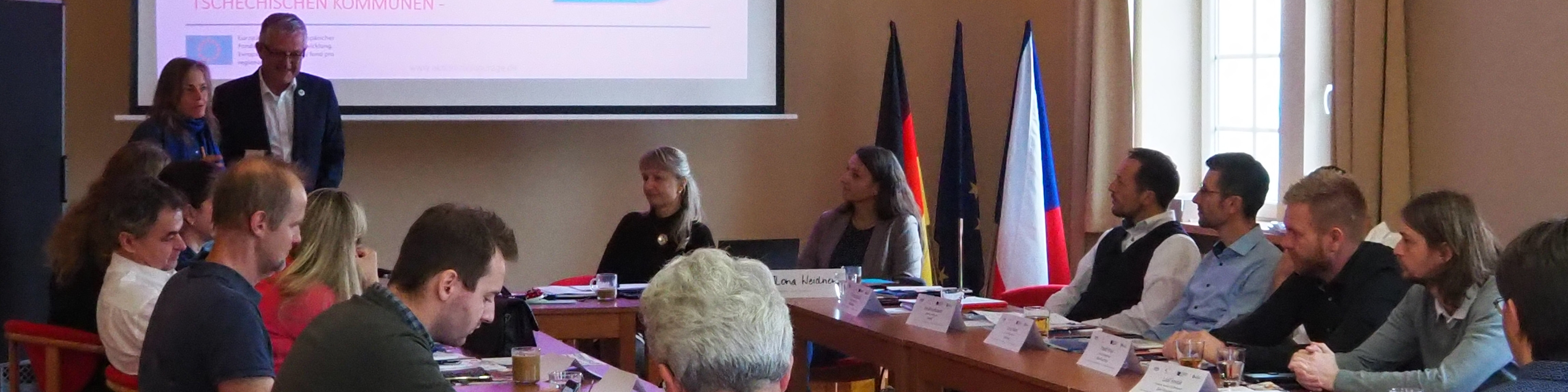 KI generiert: Das Bild zeigt eine formelle Sitzung oder Konferenz in einem Raum mit mehreren Personen, die an einem langen Tisch sitzen. Im Hintergrund sind zwei Flaggen, die deutsche und die tschechische, sowie eine Person, die vorne steht und möglicherweise eine Präsentation hält.