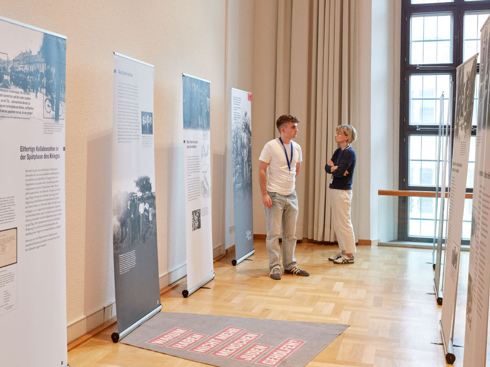 KI generiert: Das Bild zeigt eine Ausstellung in einem hellen Raum mit mehreren Informationsständern, die historische Fotos und Texte präsentieren. Zwei Personen, ein Mann und eine Frau, betrachten die Ausstellungsstücke und scheinen sich darüber auszutauschen.