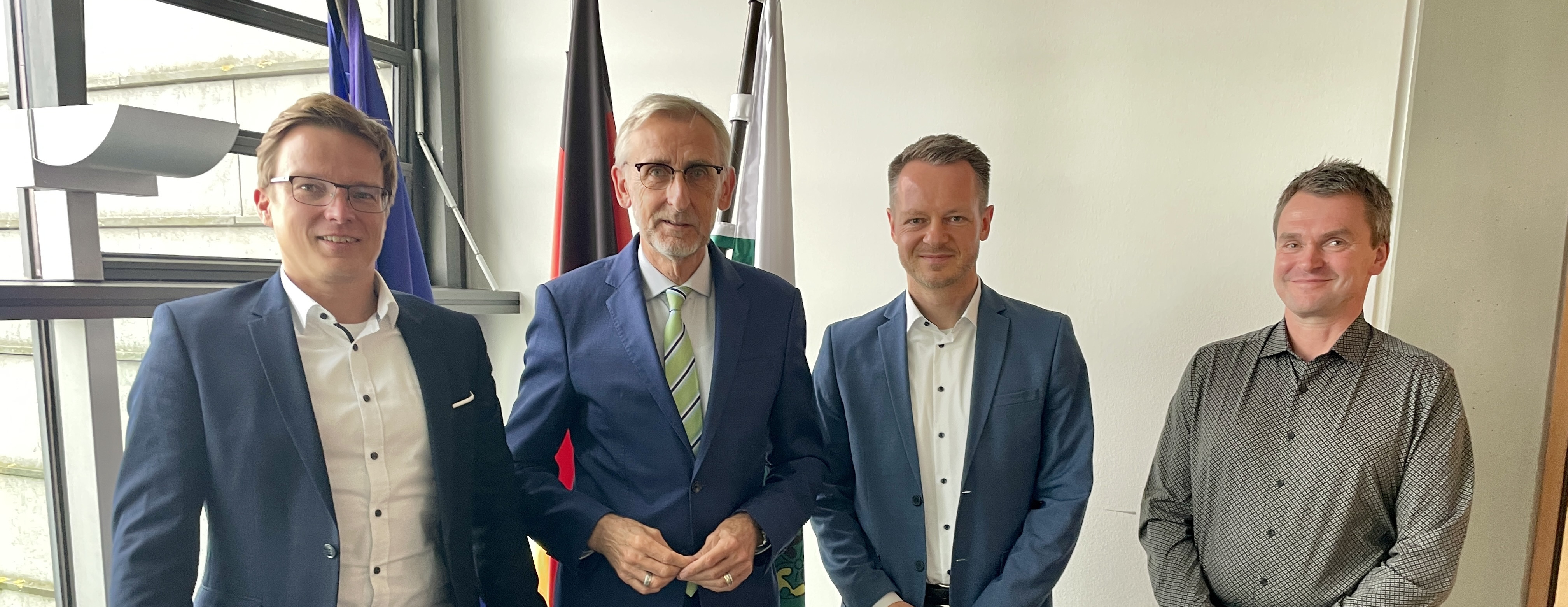 KI generiert: Das Bild zeigt vier Männer in einem Büro oder offiziellen Raum, die sich für ein Gruppenfoto versammelt haben. Im Hintergrund sind mehrere Flaggen zu sehen, darunter die deutsche Flagge.