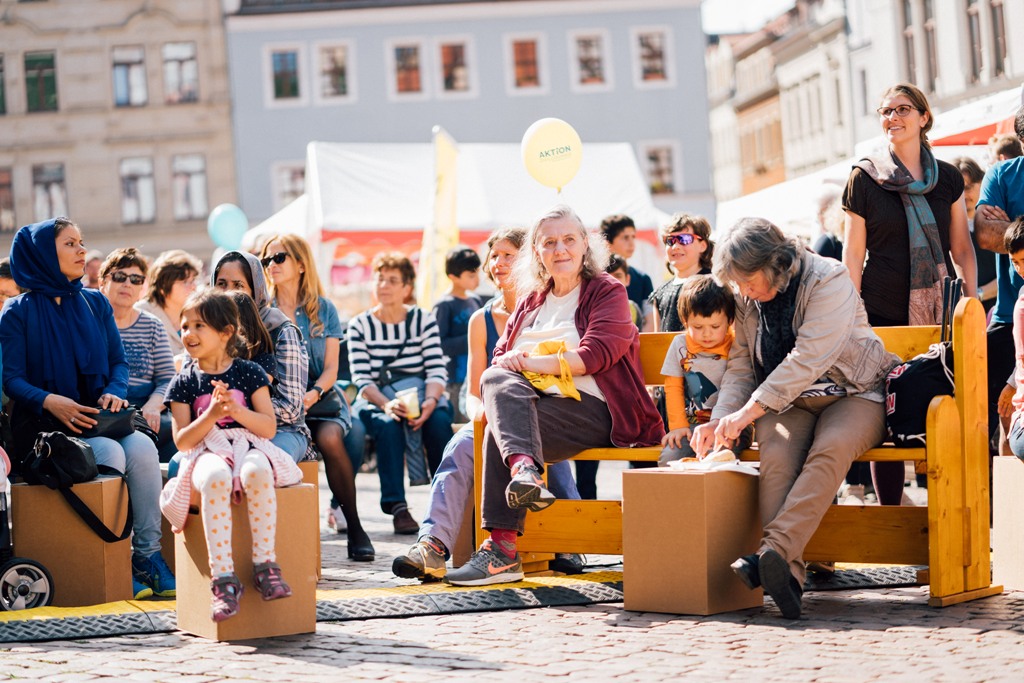 KI generiert: Das Bild zeigt eine Gruppe von Menschen, die auf provisonischen Sitzgelegenheiten aus Holz und Kartons in einer öffentlichen Platzsituation sitzen. Der Hauptinhalt des Bildes ist eine Gemeinschaftsveranstaltung oder ein Event, bei dem Menschen verschiedenen Alters zusammenkommen, um gemeinsam Zeit zu verbringen.