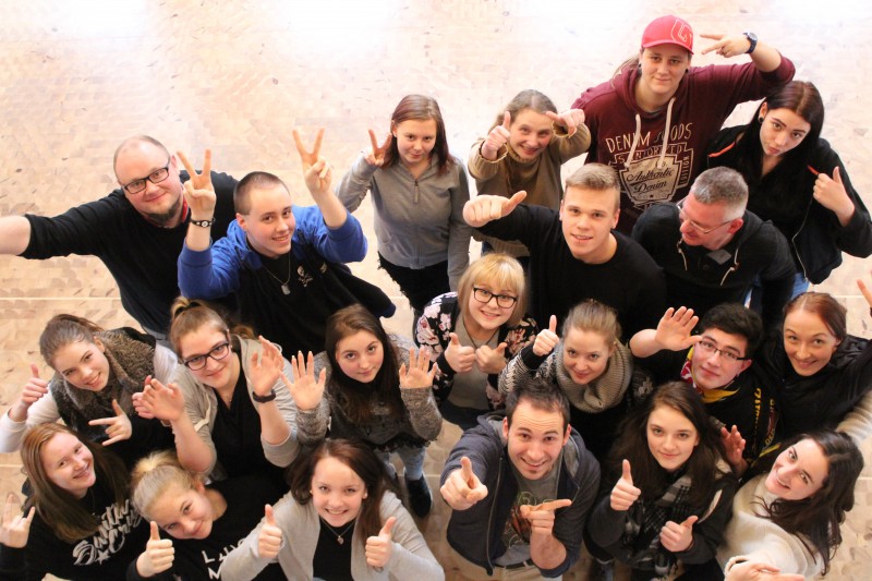 KI generiert: Das Bild zeigt eine Gruppe von etwa 20 Personen, die gemeinsam posieren und verschiedene Handgesten wie Daumen hoch, Peace-Zeichen und andere Zeichen machen. Der Hauptinhalt des Bildes ist die fröhliche und gesellige Stimmung der Gruppe.