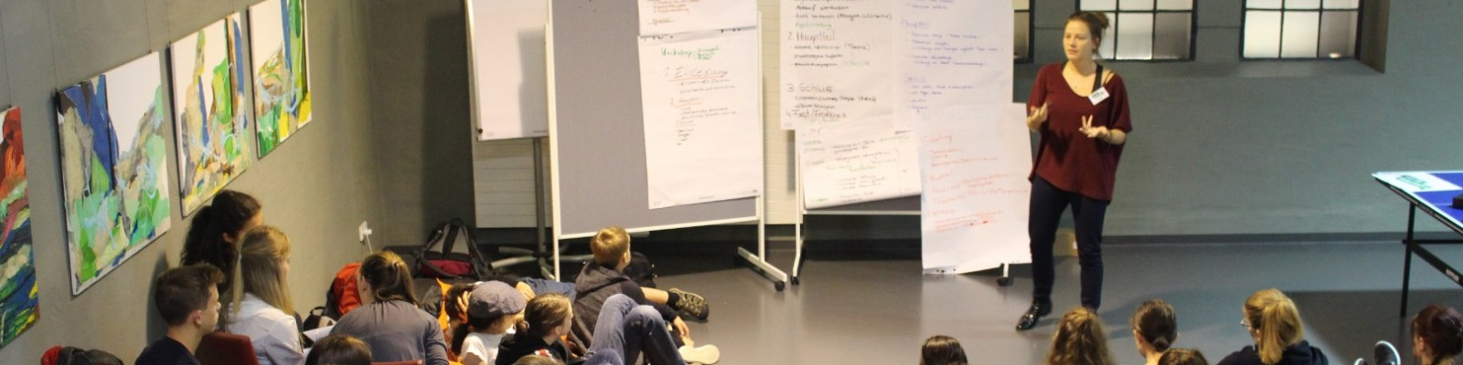 KI generiert: Das Bild zeigt eine Person, die vor einer Gruppe von sitzenden Zuhörern steht und eine Präsentation hält. Im Hintergrund sind Flipcharts mit Notizen und einige bunte Bilder an der Wand zu sehen.