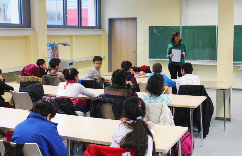 KI generiert: Das Bild zeigt eine Schulklasse, in der eine Lehrerin vor einer Gruppe von Schülern steht und etwas erklärt. Die Schüler sitzen an Tischen und scheinen aufmerksam zuzuhören.