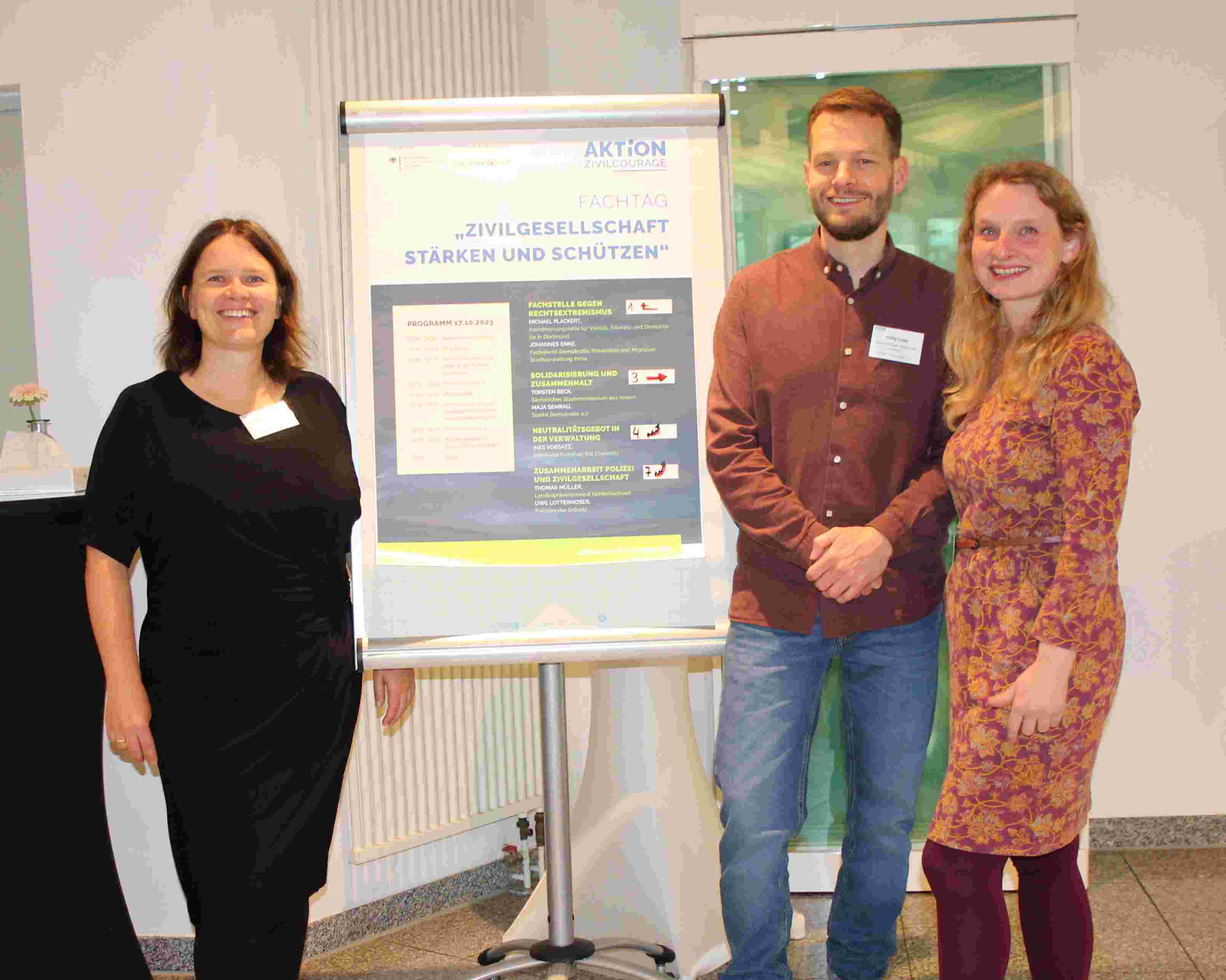 KI generiert: Das Bild zeigt drei Personen, die vor einem Flipchart mit dem Titel "Aktionstag: Zivilgesellschaft stärken und schützen" stehen. Der Hauptinhalt des Bildes ist die Präsentation oder Diskussion eines Programms oder einer Veranstaltung zu diesem Thema.