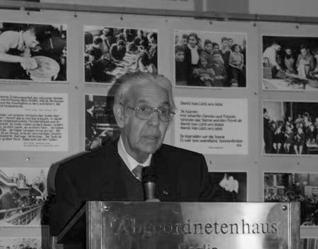 KI generiert: Das Bild zeigt einen älteren Mann mit Brille, der an einem Pult steht und spricht, während im Hintergrund eine Ausstellung mit Fotos und Texten zu sehen ist. Der Hauptinhalt des Bildes ist eine Rede oder Präsentation in einem Ausstellungsraum.