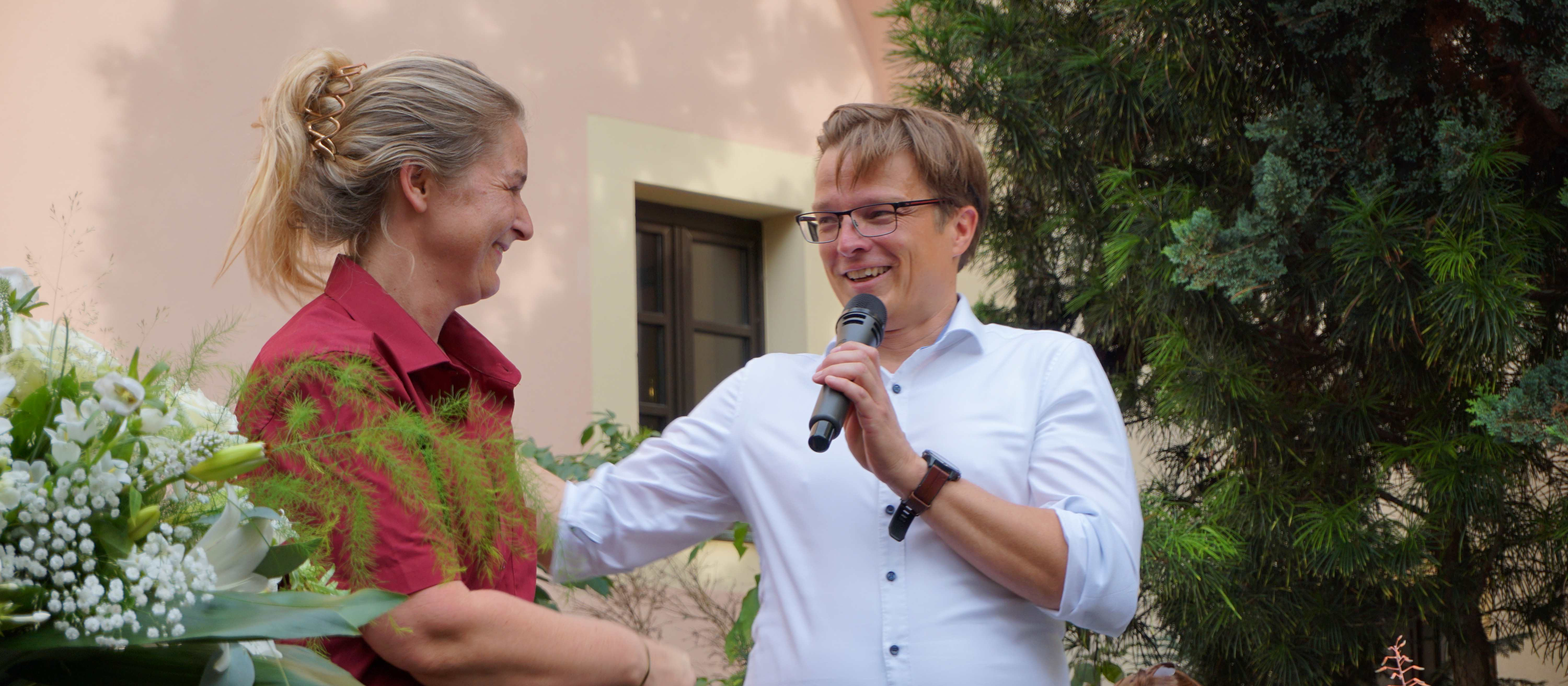 KI generiert: Das Bild zeigt eine Frau, die einem Mann einen Blumenstrauß überreicht, während der Mann ein Mikrofon hält und lächelnd mit ihr interagiert. Der Hauptinhalt des Bildes ist die freundliche und herzliche Interaktion zwischen den beiden Personen in einer scheinbar offiziellen oder feierlichen Umgebung.