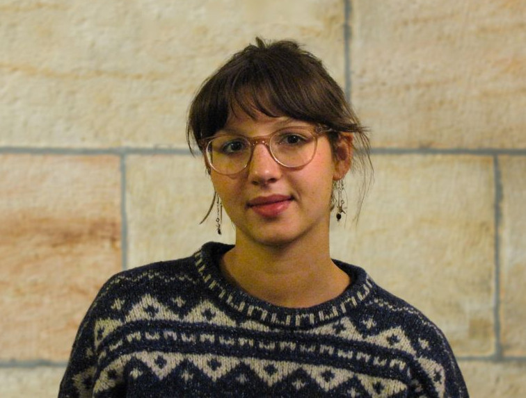 KI generiert: Das Bild zeigt eine Person mit Brille und Ohrringen, die einen dunklen Pullover mit einem geometrischen Muster trägt. Die Person steht vor einer hellen Steinwand.