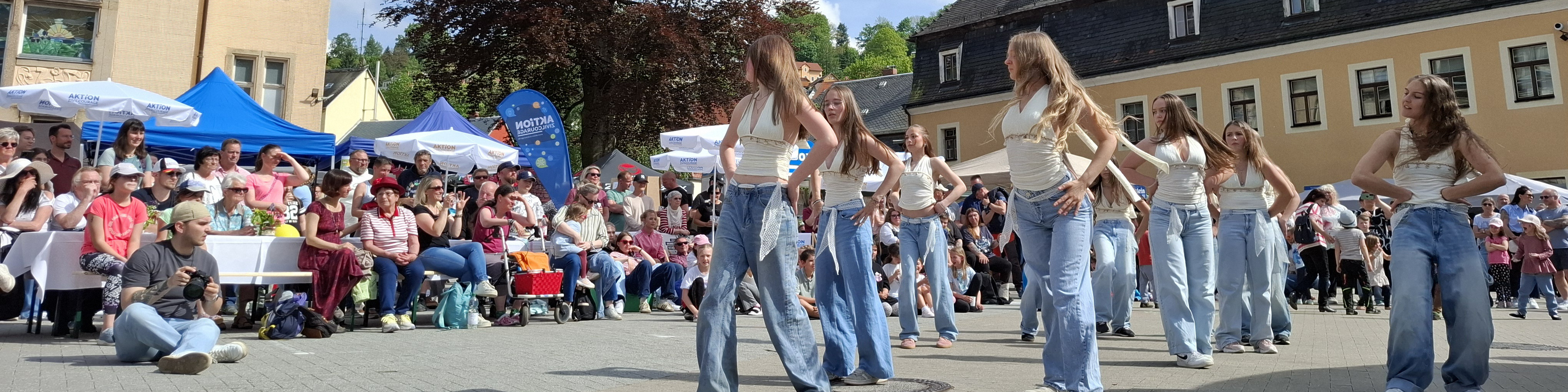 KI generiert: Das Bild zeigt eine Gruppe junger Frauen, die in einer Freiluftveranstaltung eine choreografierte Tanzdarbietung aufführen. Im Hintergrund sitzt ein Publikum, das die Darstellung beobachtet, während im Hintergrund bunte Zelte und ein Gebäude zu sehen sind.