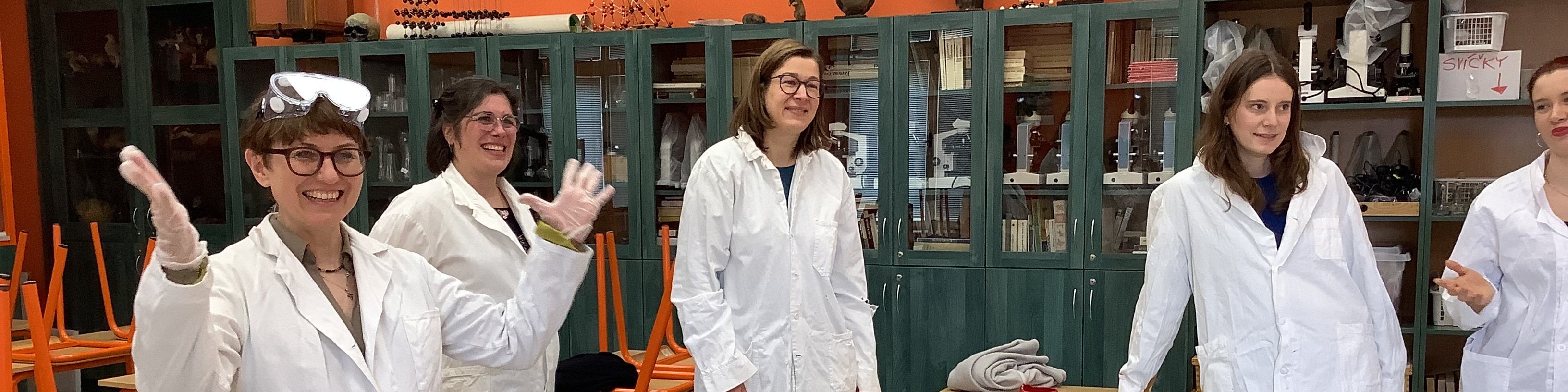 KI generiert: Das Bild zeigt fünf Personen in weißen Laborkitteln, die in einem Labor stehen und fröhlich in die Kamera winken. Im Hintergrund sind Laborgeräte, Regale mit Chemikalien und andere typische Laborausstattungen zu sehen.