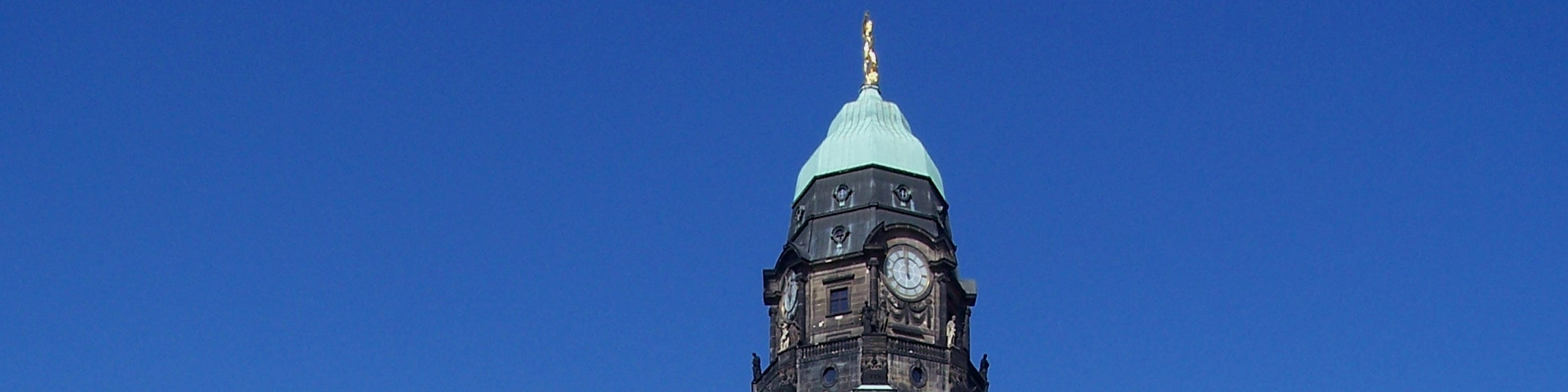 KI generiert: Das Bild zeigt den oberen Teil eines historischen Uhrturms mit einer grünen Kuppel und einer goldenen Spitze vor einem klaren blauen Himmel. Der Hauptinhalt des Bildes ist der Uhrturm.