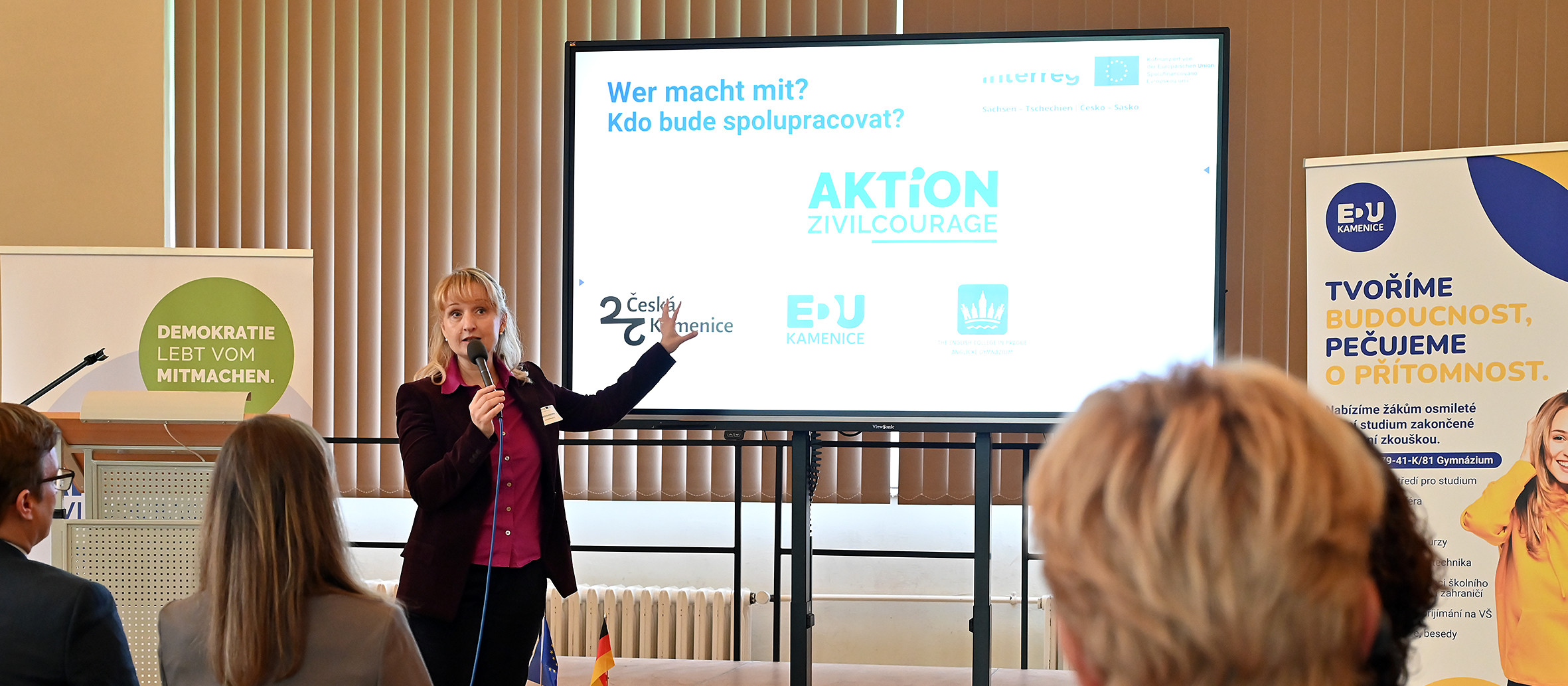 KI generiert: Das Bild zeigt eine Frau, die vor einem Publikum spricht und dabei eine Präsentation auf einer Leinwand nutzt. Der Hauptinhalt der Präsentation scheint die Frage "Wer macht mit? Kdo bude spolupracovat?" (Wer macht mit? Wer wird zusammenarbeiten?) im Rahmen der "AKTION Zivilcourage" zu sein.