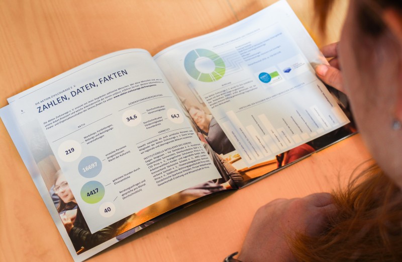 KI generiert: Das Bild zeigt eine Person, die ein aufgeschlagenes Buch oder eine Broschüre mit Diagrammen, Zahlen und Texten auf Deutsch liest. Der Hauptinhalt scheint eine statistische oder analytische Darstellung zu sein, die verschiedene Daten und Fakten enthält.