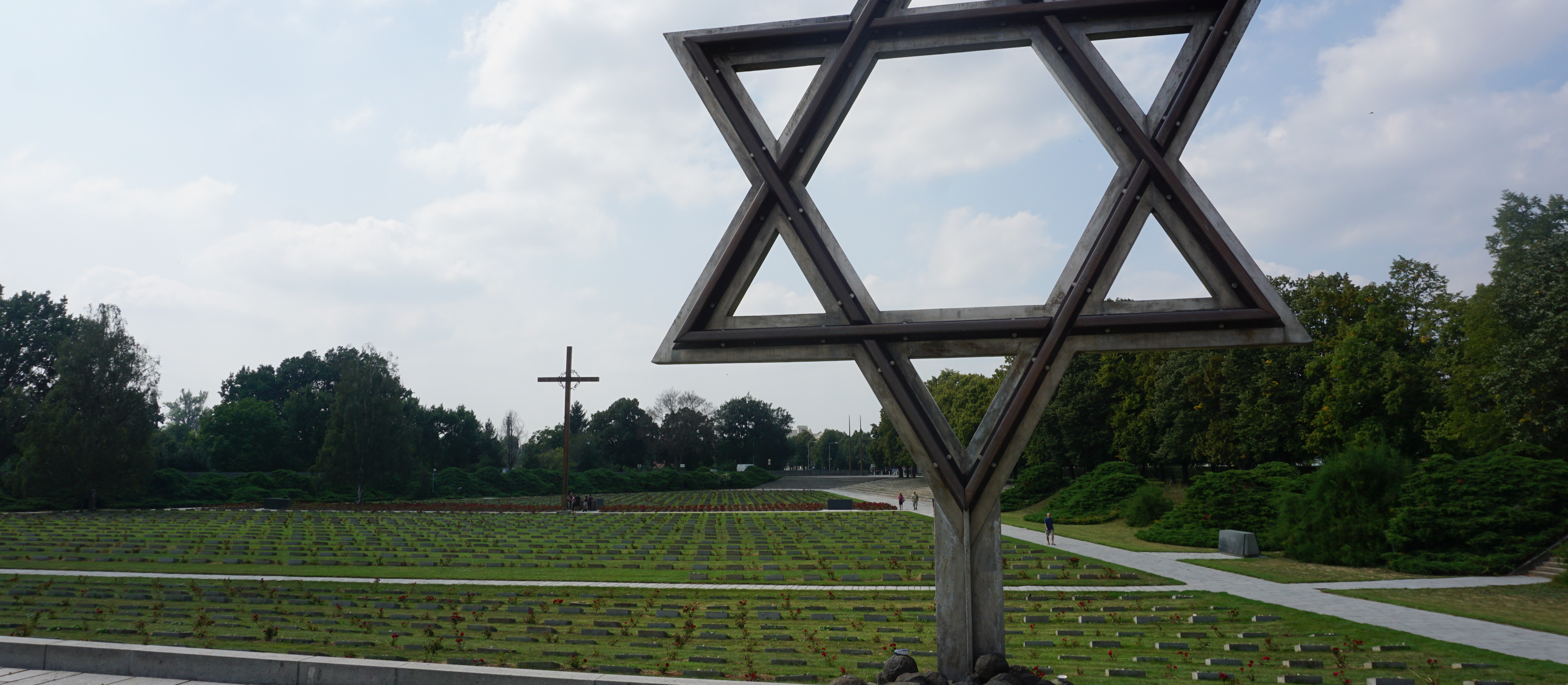 KI generiert: Das Bild zeigt einen großen Davidstern in einem weitläufigen, gepflegten Park mit Reihen von Grabsteinen oder Gedenktafeln. Im Hintergrund sind ein Kreuz und eine Baumreihe zu sehen, was auf einen interreligiösen Gedenkort oder Friedhof hindeutet.