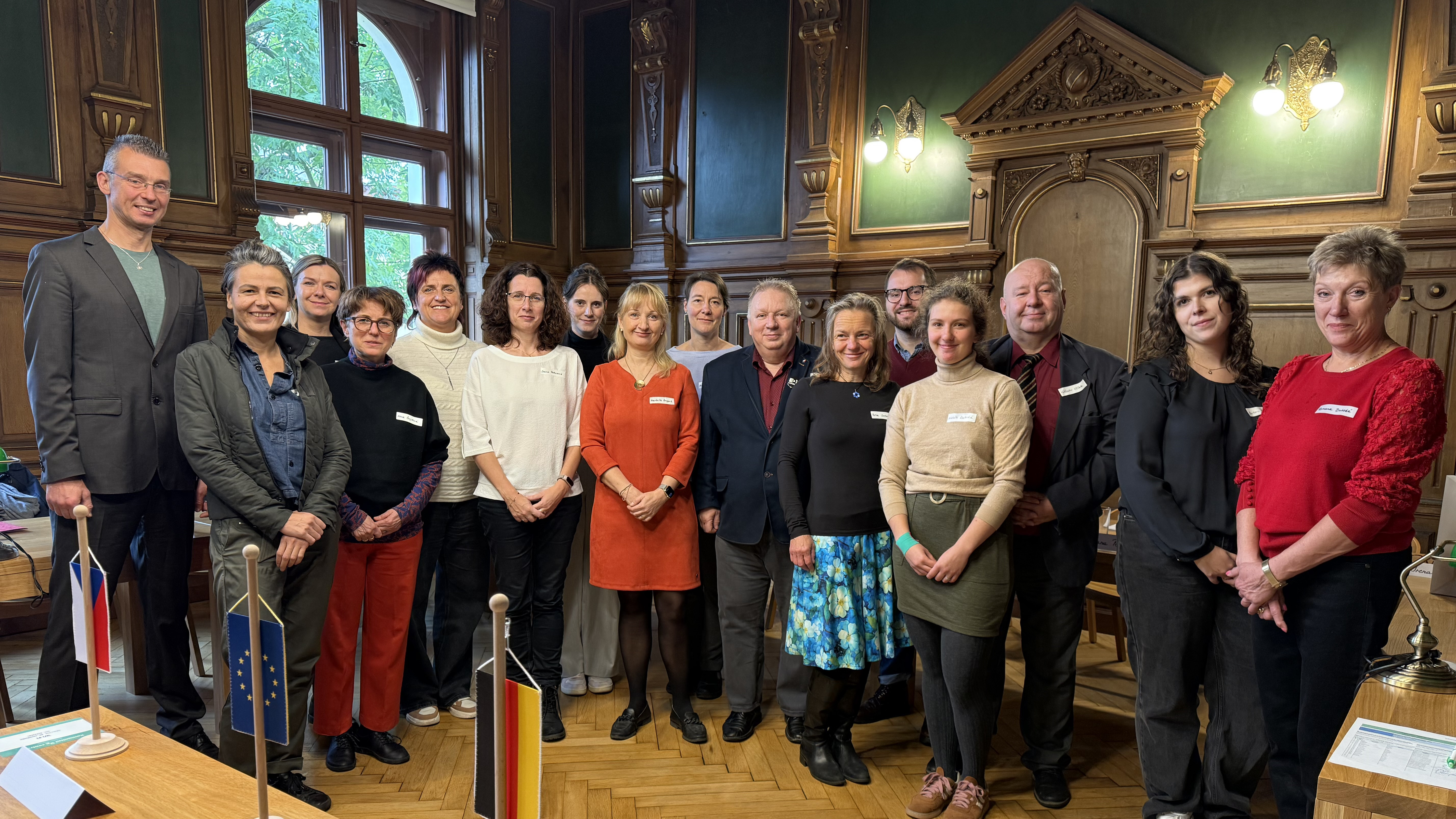 KI generiert: Das Bild zeigt eine Gruppe von Personen, die in einem historischen Raum mit Holzvertäfelung und großen Fenstern stehen. Im Vordergrund sind drei kleine Flaggen zu sehen: die deutsche Flagge, die Flagge der Europäischen Union und eine weitere, nicht klar erkennbare Flagge.