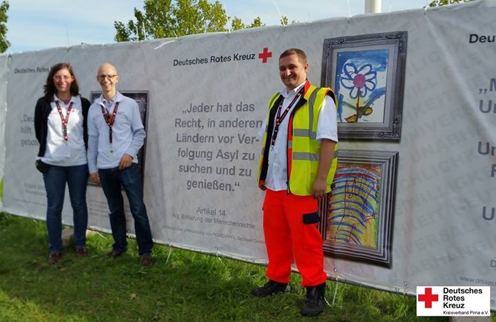 KI generiert: Das Bild zeigt drei Personen vor einem Banner des Deutschen Roten Kreuzes, das sich für die Rechte von Asylsuchenden einsetzt. Der Hauptinhalt des Bildes ist die Unterstützung und Förderung der Menschenrechte durch das Deutsche Rote Kreuz.