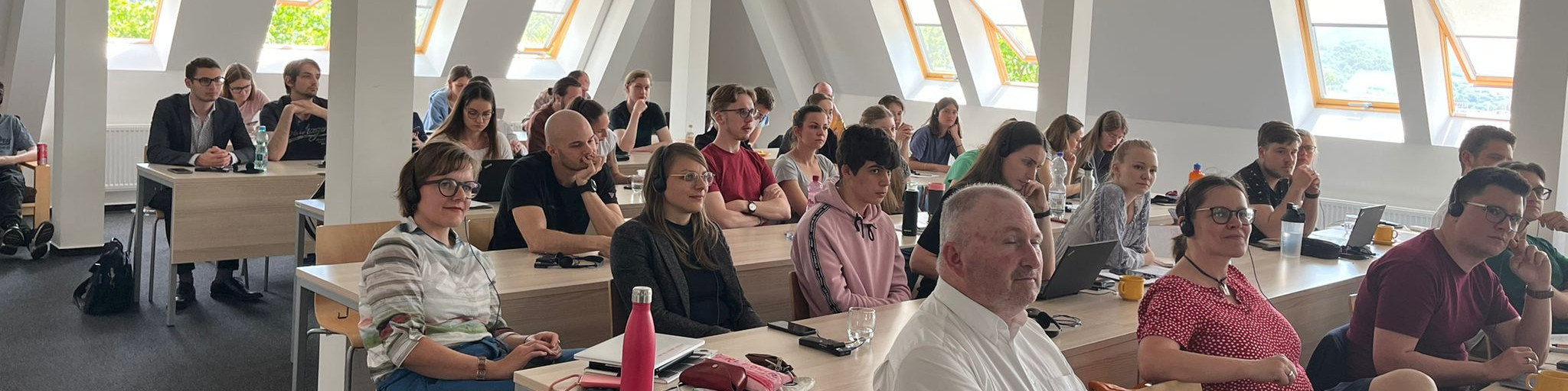 KI generiert: Das Bild zeigt eine Gruppe von Personen, die in einem hellen Raum mit großen Fenstern an langen Tischen sitzen und scheinbar einer Präsentation oder einem Vortrag folgen. Der Hauptinhalt des Bildes ist eine Versammlung oder ein Seminar in einem modernen, gut beleuchteten Innenraum.