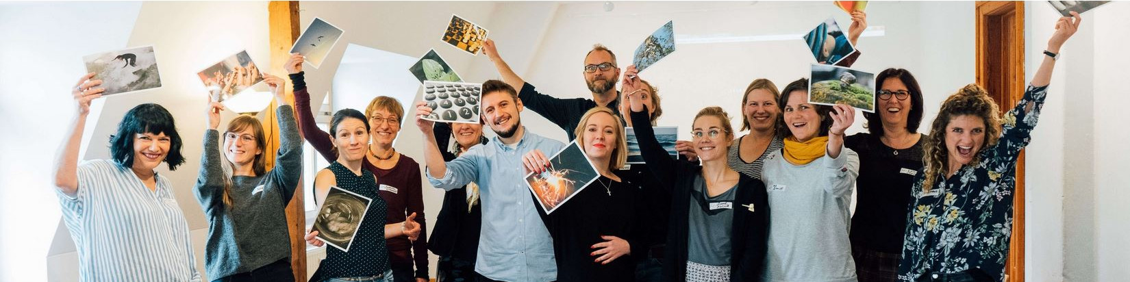 KI generiert: Das Bild zeigt eine Gruppe von Menschen, die fröhlich in die Kamera lächeln und Fotos in die Luft halten. Der Hauptinhalt des Bildes ist eine Gruppe von Personen, die gemeinsam eine positive und enthusiastische Stimmung teilen, möglicherweise bei einem Workshop oder einer Veranstaltung.