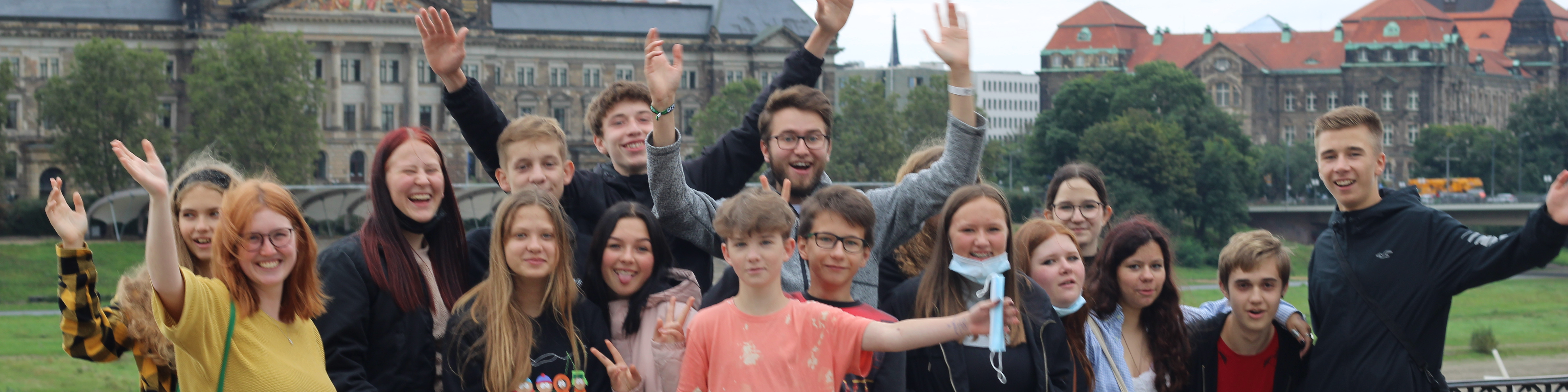 KI generiert: Das Bild zeigt eine Gruppe von Jugendlichen und jungen Erwachsenen, die draußen vor einer historischen Kulisse mit Gebäuden und Grünflächen posieren und dabei fröhlich in die Kamera winken. Der Hauptinhalt des Bildes ist die lebhafte und freundliche Stimmung der Gruppe in einer urbanen Umgebung.