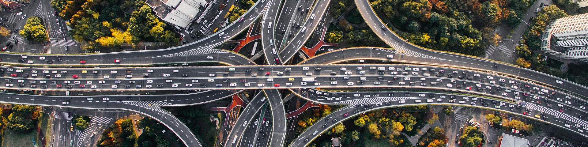 KI generiert: Das Bild zeigt eine komplexe, mehrspurige Autobahnkreuzung aus der Vogelperspektive mit dichtem Verkehr. Der Hauptinhalt des Bildes ist das verzweigte Netz aus Straßen und Brücken, umgeben von einigen grünen Baumkronen und Gebäuden.