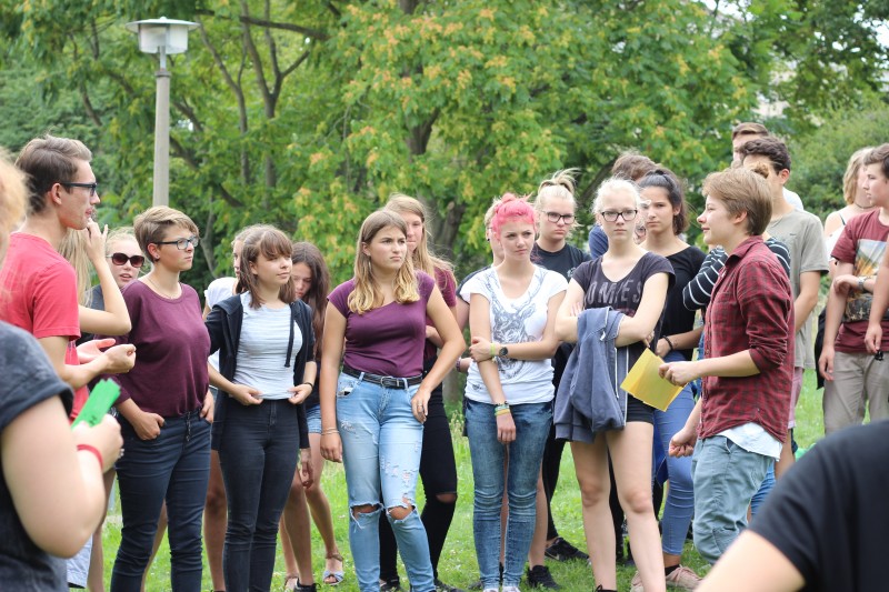 KI generiert: Das Bild zeigt eine Gruppe junger Menschen, die im Freien stehen und sich unterhalten. Der Hauptinhalt des Bildes ist eine gesellige Zusammenkunft in einem parkähnlichen Umfeld.